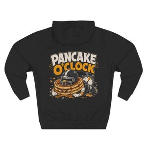 May include: A black hooded sweatshirt featuring a graphic of a stack of pancakes wearing a football helmet, with the text PANCAKE O'CLOCK above it. The design includes a stopwatch and syrup drips, rendered in shades of yellow, white, and black.