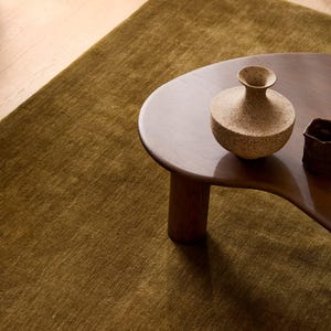 May include: A dark olive green area rug with a short pile texture, featuring a dark brown wooden coffee table with a curved edge. On the table sits a speckled beige ceramic vase with a narrow neck and a small dark brown ceramic vessel.