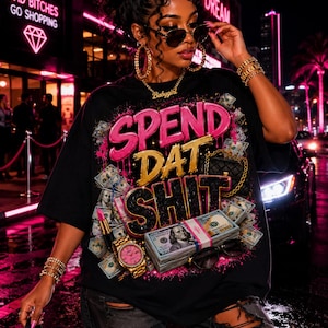 May include: A black short-sleeve t-shirt featuring a graphic print with the text SPEND DAT SHIT in pink, gold, and black. The design includes images of stacks of hundred-dollar bills, a gold watch, a lipstick tube, and a small quilted handbag.