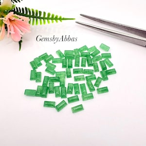 May include: A collection of small, rectangular, translucent green gemstones scattered across a white surface. The text GemsbyAbbas is printed in black. A pair of silver metal tweezers and a pink and green artificial flower are visible.