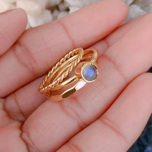 Puede incluir: Un conjunto de tres anillos dorados apilados. Un anillo presenta una textura de cuerda trenzada, otro es una banda lisa y sencilla, y el tercero es una banda martillada rematada con un pequeño engaste de piedra labradorita azul iridiscente.