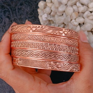 May include: A set of five copper-colored metal bangles stacked together. Each bangle features a distinct embossed pattern, including floral motifs, geometric shapes, and braided textures. The bracelets have a metallic finish and a uniform width.