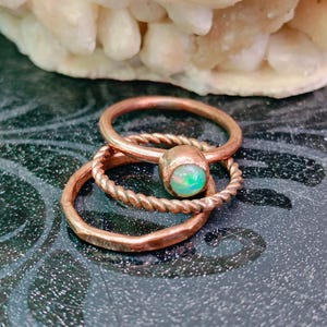Könnte beinhalten: Ein Set aus drei kupferfarbenen Ringen, die übereinander gestapelt sind. Ein Ring hat eine glatte Schiene, einer eine gedrehte Kordelstruktur und der dritte ist ein schlichter Ring mit einem kleinen, runden, irisierenden Opal-Cabochon.