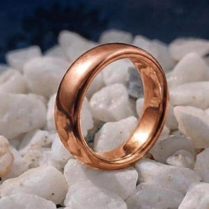 May include: A polished, smooth, solid copper-coloured ring with a rounded, domed profile. The band features a continuous, seamless design with a high-shine metallic finish that reflects light across its curved surface, resting upright.
