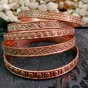 May include: A stack of five copper-coloured metal bangles, each featuring a distinct embossed pattern. The designs include geometric squares, floral motifs, repeating four-petaled shapes, and a series of raised circular dots along the bands.