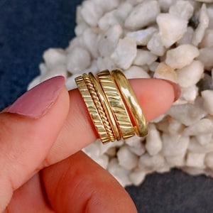 May include: A set of four gold-toned rings worn together on a finger. The stack includes one ring with a twisted rope texture, one with diagonal etched lines, and two smooth, plain bands, all featuring a polished metallic finish.