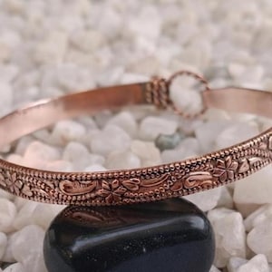 Può includere: Un bracciale rigido in metallo color rame con un motivo floreale e a viticci in rilievo sulla superficie esterna. Il bracciale presenta un design circolare con un piccolo meccanismo di chiusura rotondo integrato a un'estremità.