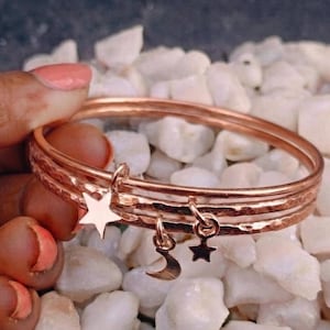 May include: A set of three rose gold-toned metal bangles with a hammered texture. The bracelets feature three dangling charms: a large five-pointed star, a small crescent moon, and a small five-pointed star, all in a matching rose gold-toned metal.