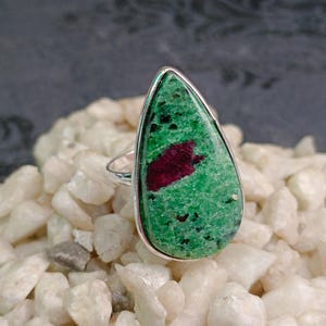 Può includere: Un anello a forma di goccia con una pietra di zoisite verde lucida caratterizzata da un'evidente inclusione di rubino color magenta intenso. La gemma è incastonata in una semplice fascia in metallo argentato che segue il profilo affusolato.