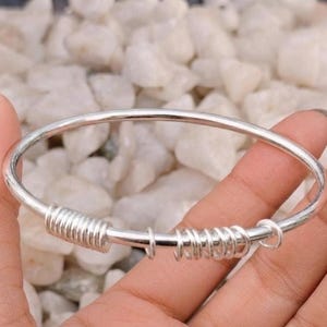 May include: A silver-toned bangle bracelet featuring a smooth, circular band adorned with two distinct sections of coiled wire detailing and several small, loose silver-toned rings that slide along the main body of the bracelet.