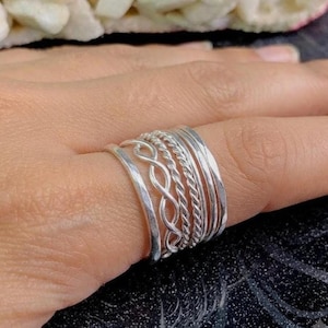 Puede incluir: Un conjunto de cinco anillos de plata apilados en un dedo. La colección incluye una banda lisa pulida, una banda trenzada estilo cuerda, una banda trenzada estilo infinito y dos bandas de plata finas y sencillas con acabado metálico brillante.