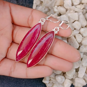 May include: A pair of long, marquise-shaped earrings featuring deep magenta-red stones set in polished silver-toned metal frames. The earrings are designed with secure lever-back closures, showcasing a smooth, elongated silhouette and vibrant colour.