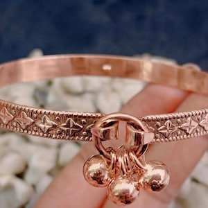 Può includere: Un bracciale rigido in metallo color oro rosa caratterizzato da un motivo a stelle in rilievo ripetuto lungo la fascia. Il bracciale presenta un anello centrale da cui pendono tre piccoli campanelli rotondi in metallo lucido.