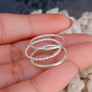 Puede incluir: Un conjunto de tres anillos de plata con diferentes texturas de banda. Un anillo tiene un acabado pulido liso, otro tiene un diseño de cuerda trenzada y otro tiene una superficie martillada. Los tres anillos están apilados en un diseño simple.