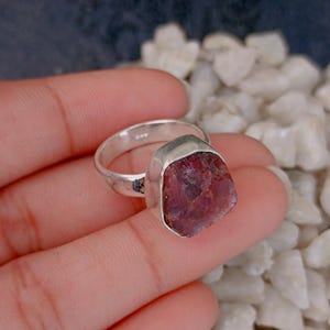 Puede incluir: Un anillo de plata con una gran piedra preciosa en bruto y sin tallar, de un tono rojo púrpura intenso. La piedra está engastada en un sencillo engaste de plata pulida que resalta la textura natural e irregular del mineral.