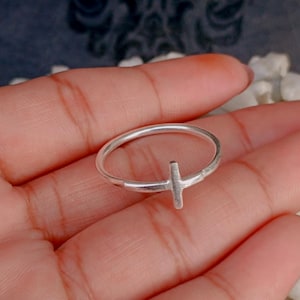 Peut inclure: Une fine bague en argent poli présentant un petit élément en forme de croix verticale intégré à l'anneau. La bague possède une finition métallique lisse et une forme circulaire simple, mettant en valeur un design épuré avec une croix.
