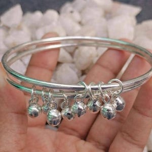 May include: A silver bangle bracelet featuring two thin, smooth bands connected together. The bracelet is adorned with a series of small, round, polished silver bell charms that hang from the bands, creating a delicate and reflective metallic appearance.