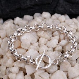 Könnte beinhalten: Ein silbernes Gliederarmband mit polierter Oberfläche und Knebelverschluss. Das Armband besteht aus einer Reihe miteinander verbundener kreisförmiger Metallglieder, wobei ein runder Ring und ein T-Stück den sicheren Verschluss bilden.