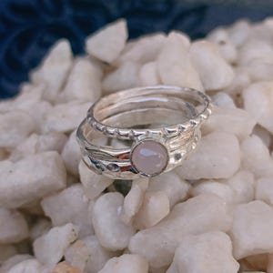 Può includere: Un set di tre anelli in argento impilati con finitura lucida. Un anello è liscio, uno ha una superficie martellata testurizzata e il terzo anello è sormontato da una piccola pietra cabochon rotonda rosa pallido incastonata in una montatura d'argento.