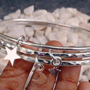 May include: A set of three silver-toned bangles featuring a hammered texture. The bracelets are adorned with three dangling charms: a large five-pointed star, a smaller five-pointed star, and a crescent moon charm, all in a polished silver finish.