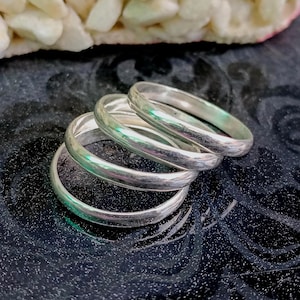 Può includere: Un set di quattro anelli color argento lucido con un profilo a fascia liscio e arrotondato. Gli anelli sono impilati in modo leggermente sovrapposto, mostrando una superficie metallica riflettente e un design circolare semplice e uniforme.