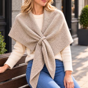 May include: A beige heathered knit shawl draped over the shoulders and tied in a knot at the front, worn over a cream-colored long-sleeve ribbed sweater and blue denim jeans. The shawl features a textured wool-like appearance.