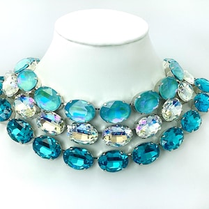 May include: A statement necklace featuring three rows of large, oval-shaped turquoise and iridescent crystal stones. The necklace is displayed on a white mannequin bust, showcasing its bold design and color palette.
