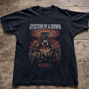 Puede incluir: Una camiseta negra de manga corta con un estampado gráfico que incluye el texto SYSTEM OF A DOWN y AERIALS. El diseño muestra un ojo central con tentáculos mecánicos sobre un horizonte urbano bajo una nube de color naranja y rojo.