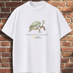 May include: A white short-sleeve t-shirt featuring a graphic of a turtle in the centre. Below the turtle, the text reads "slow motion over no motion" followed by the National Geographic logo with its signature yellow rectangular border.