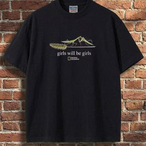 May include: A black short-sleeve t-shirt featuring a graphic of two green praying mantises on a plant stem. Below the illustration, the text reads "girls will be girls" in white, followed by the National Geographic logo in a yellow rectangle.