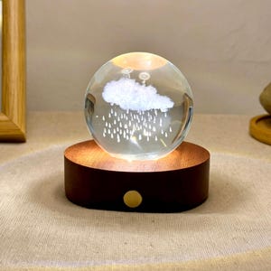 May include: A clear glass sphere containing a white etched cloud and rain design, illuminated from below by a circular dark wood base with a small gold-colored button on the front. The light casts a warm glow upward through the glass orb.