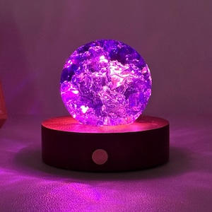 May include: A spherical glass orb with a textured, cracked interior surface illuminated by a vibrant purple light. The orb rests on a circular, dark brown wooden base featuring a small, centred circular button on its front side.