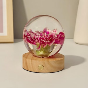 Peut inclure: Un presse-papier sphérique transparent contenant une fleur d'œillet rose et blanche, monté sur une base circulaire en bois clair avec un petit bouton métallique centré sur la face avant du bois.