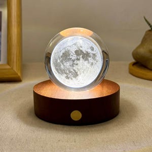 May include: A clear glass sphere featuring a detailed 3D laser-engraved moon inside, resting on a circular dark brown wooden base. The base includes a small, centered gold-colored circular button on the front, providing a warm light to illuminate the moon.