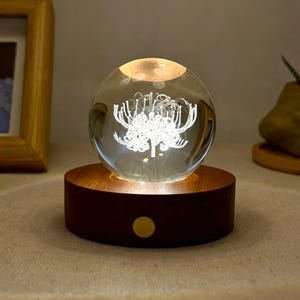 May include: A clear glass sphere lamp featuring an internal laser-etched white spider lily design. The sphere rests on a circular dark brown wooden base with a small gold-colored circular button on the front, emitting a warm light from below.