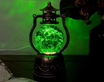 Könnte beinhalten: Eine Laternenlampe mit einem dunkelbronzefarbenen Metallrahmen, der an der Basis mit Schnörkeln verziert ist, und einer zentralen kugelförmigen Glaskugel, die ein helles, leuchtend grünes Licht ausstrahlt. Das Glas hat eine texturierte Rissoptik.
