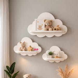 May include: Three white cloud-shaped wall shelves mounted on a gray wall. The top shelf holds a picture frame, a teddy bear, and a wooden camera. The middle shelf holds a small bear and a star. The bottom shelf holds a plant and a plush toy.