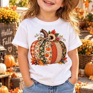 May include: A white short-sleeved t-shirt featuring a large graphic of a pumpkin composed of various patterned segments including plaid, floral, and polka dot prints, accented with a plaid bow, sunflower, and autumn leaves.