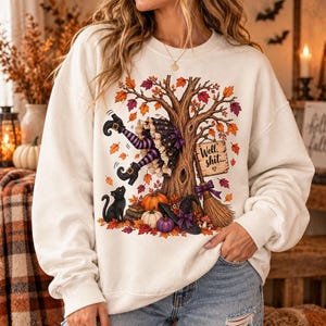 May include: A cream-colored crewneck sweatshirt featuring a graphic of a tree with autumn leaves, a witch's legs in striped stockings, a black cat, pumpkins, a broom, and a sign that reads Well, shit... with a small heart symbol below the text.