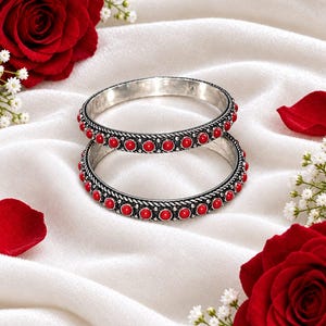 May include: Two silver bangles featuring a repeating pattern of small, round, bright red stones set along the outer circumference. The metal bands have a textured, twisted rope design bordering the row of red stones, creating a detailed finish.