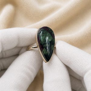 May include: A ring featuring a large, teardrop-shaped cabochon stone with a mottled pattern of deep forest green and dark magenta-red. The stone is set in a smooth, polished gold-toned metal bezel and attached to a thin, simple gold-toned band.