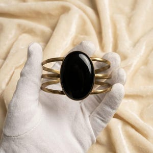 Könnte beinhalten: Ein Armreif mit einem großen, glatten, ovalen schwarzen Stein in einer messingfarbenen Metallfassung. Das Armband besteht aus drei parallelen, dünnen, geschwungenen Metalldrähten, die mit der zentralen Fassung verbunden sind.
