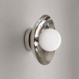 May include: A wall sconce featuring a polished silver-tone metallic bowl-shaped reflector and a central spherical white glass globe light. The fixture is mounted on a circular base and displays a reflective surface with visible light reflections.