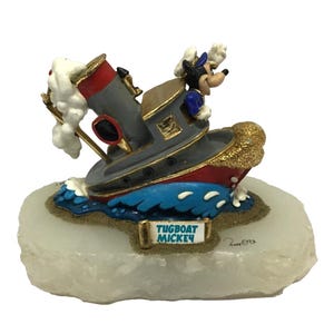 May include: A figurine of Mickey Mouse piloting a grey and red tugboat on blue waves. The boat features a red smokestack with white smoke, gold trim, and a sign reading TUGBOAT MICKEY. The entire piece is mounted on a textured white base.