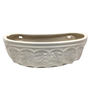 May include: A semi-circular off-white ceramic wall planter featuring a raised relief pattern of grape clusters and leaves along the exterior. The vessel has a flat back with two integrated rectangular holes for mounting against a vertical surface.