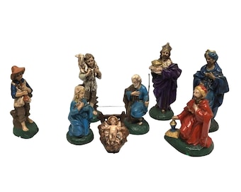 May include: A nativity set featuring eight hand-painted figurines including a central infant in a manger, kneeling figures in blue and red robes, a standing figure in purple, a shepherd with a lamb, and a standing figure in a brown hat and blue shirt.