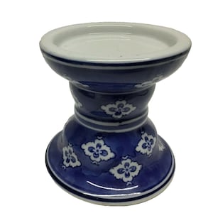 May include: A ceramic candle holder featuring a deep blue glaze with a repeating white floral pattern. The piece has a wide, circular base, a tapered middle section, and a broad, flat top rim designed to securely hold a pillar candle.