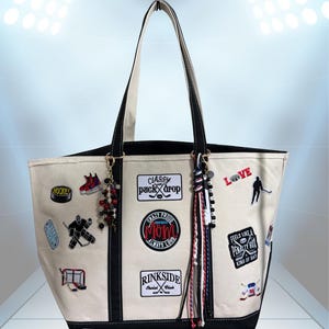 May include: A cream-colored canvas tote bag with black trim and handles, decorated with various hockey-themed embroidered patches and text including "Classy Puck Drop," "Crazy Proud Hockey Mom," and "Rinkside Social Club," plus beaded charms.