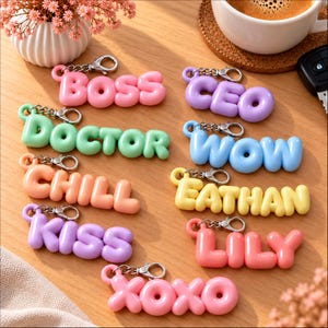 Könnte beinhalten: Eine Sammlung von neun bunten Schlüsselanhängern im Bubble-Letter-Stil mit den Wörtern BOSS, CEO, DOCTOR, WOW, CHILL, EATHAN, KISS, LILY und XOXO. Jeder Anhänger besteht aus glatten, abgerundeten Kunststoffbuchstaben mit einem silbernen Karabinerhaken.