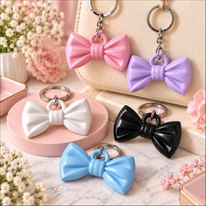 May include: A collection of five bow-shaped keychains in solid colours including pink, lavender, white, black, and light blue. Each keychain features a smooth, glossy finish, a central knot detail, and a silver-toned metal split ring attached to the top.